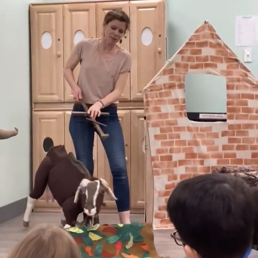 Woman with goat puppet performing in front of a toy house and an audience.