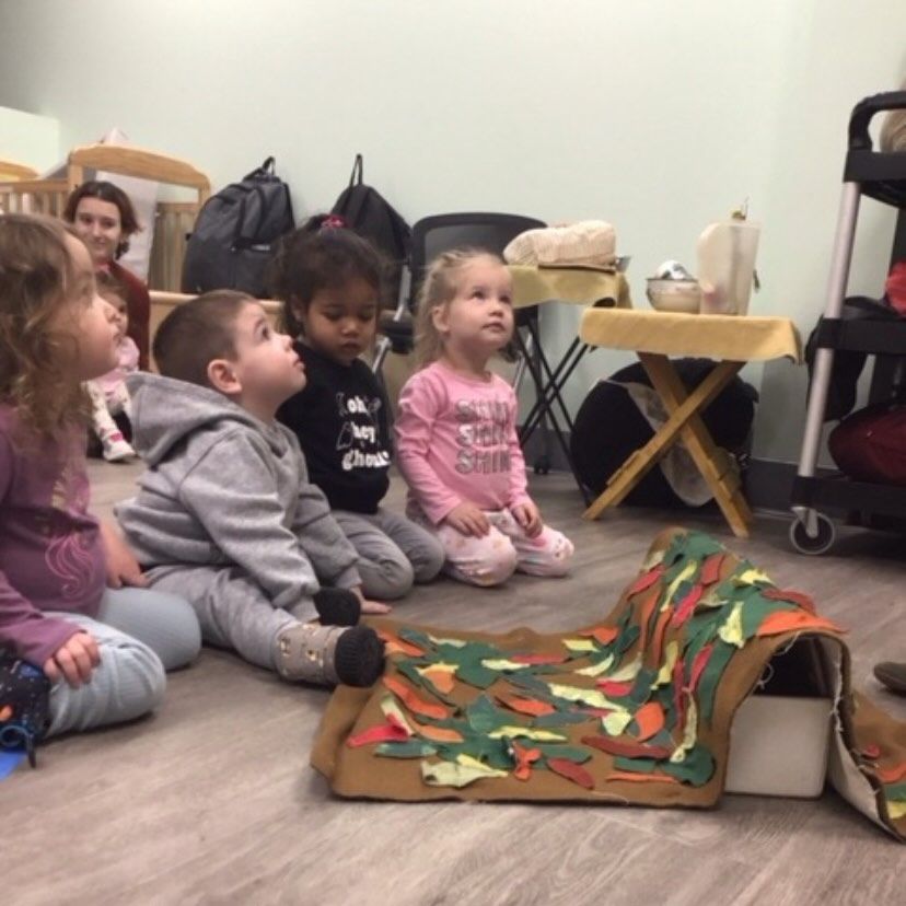 Children sit on the floor, watching a felt tunnel with colorful leaves. An adult is in the background.