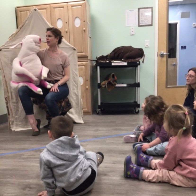 Woman with a pig puppet reads to children in a room with a tent and a rolling cart.