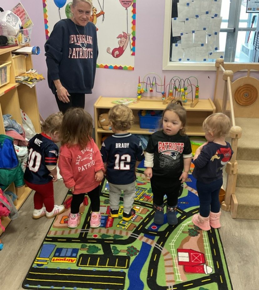 A woman and five children in a playroom. Children wear Patriots apparel, standing on a road rug.