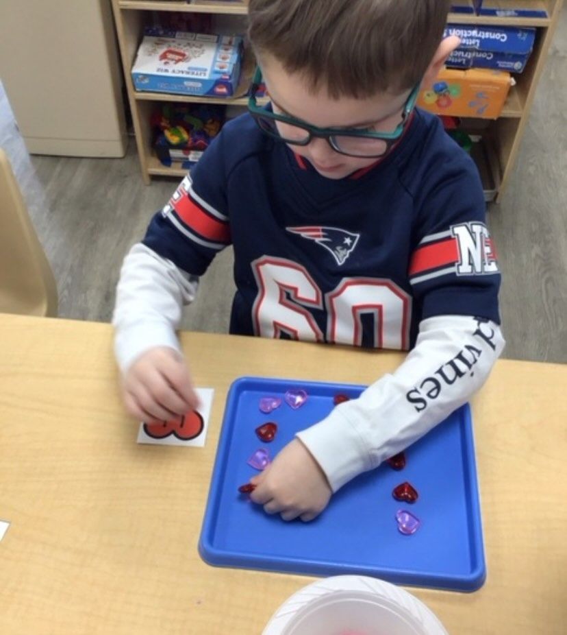A child in glasses counts heart-shaped objects on a tray while looking at a card with a number.