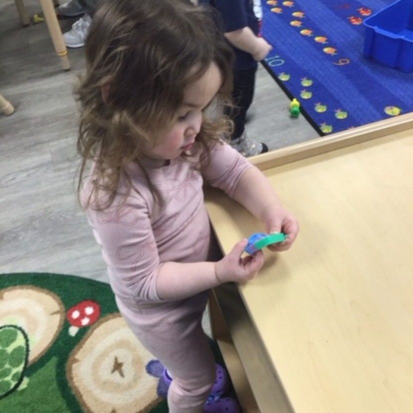 Child in pink pajamas examines toy at a table in a classroom.