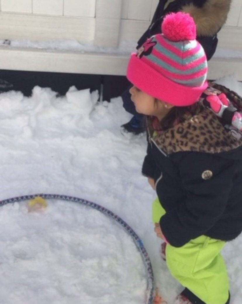 Child wearing pink hat and green pants looks at a hula hoop in the snow.
