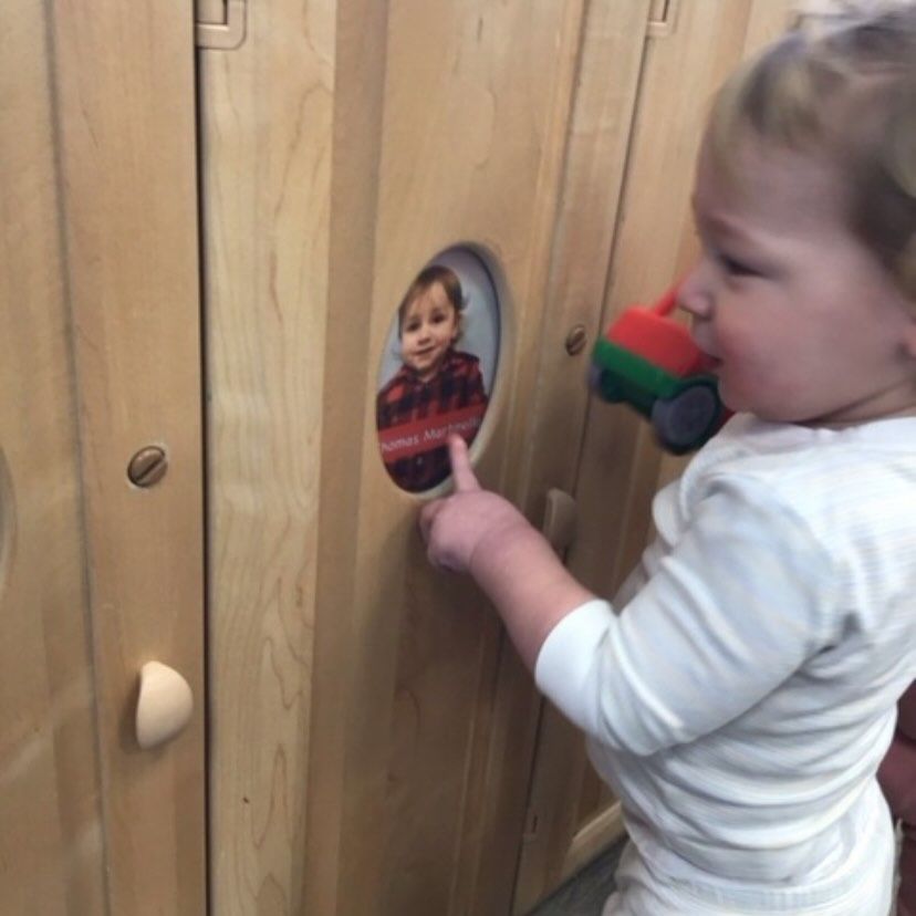 Toddler pointing at a picture in a wooden cabinet, holding a toy truck, in a play area.