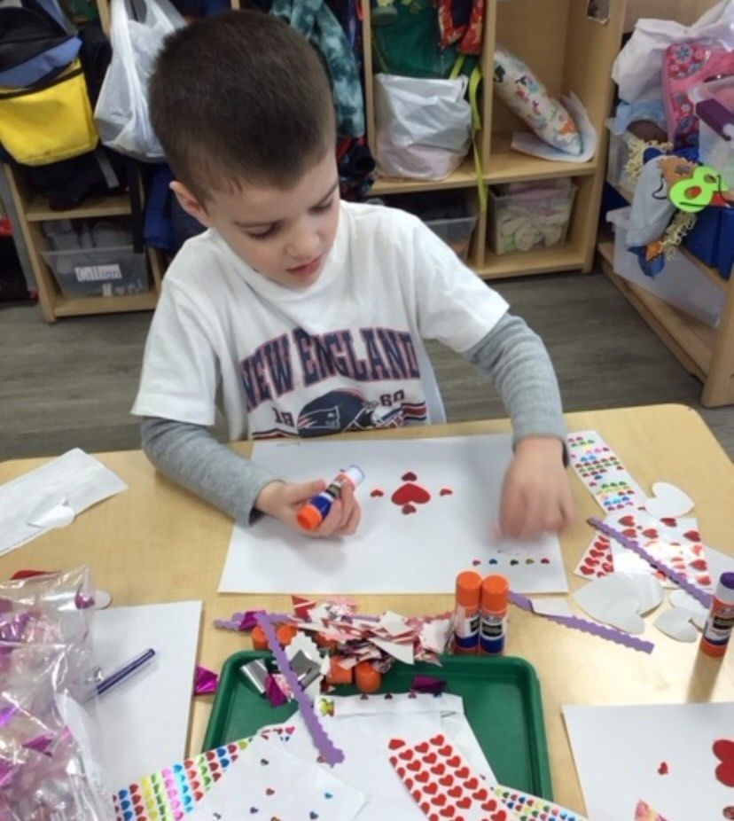 Boy in white shirt gluing hearts to paper at a table in a classroom.