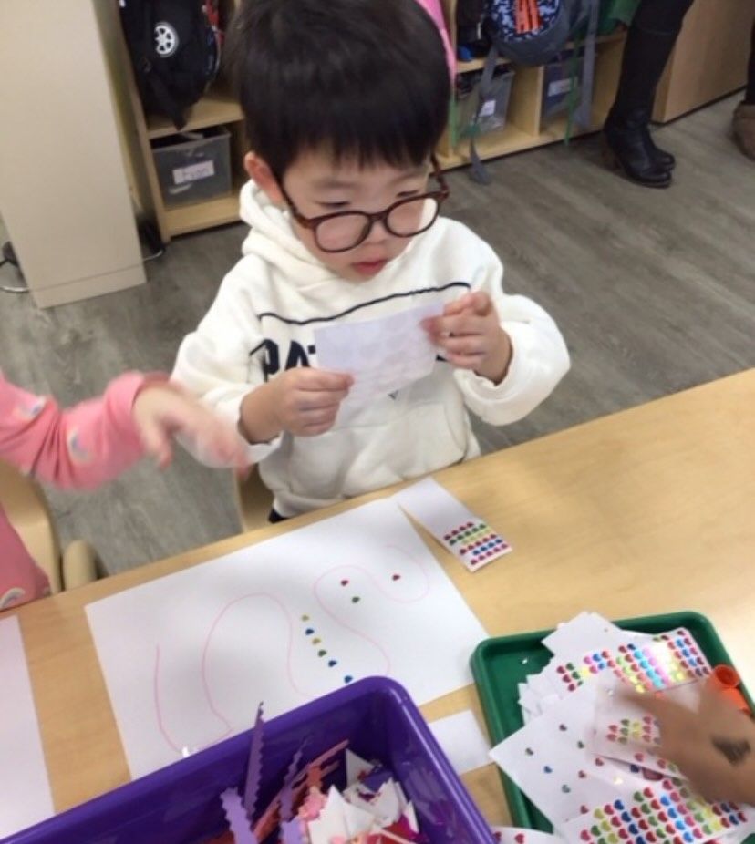 Young child with glasses examines paper while crafting at a table with stickers.