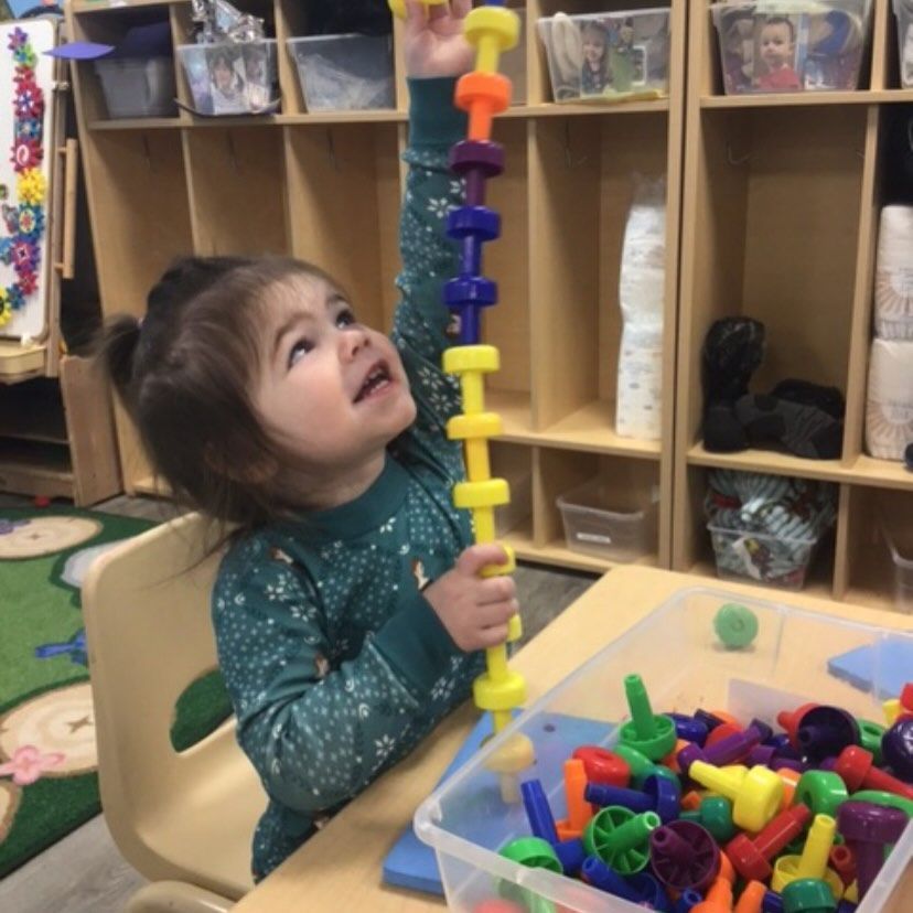 Child building a colorful tower of blocks at a table in a classroom.