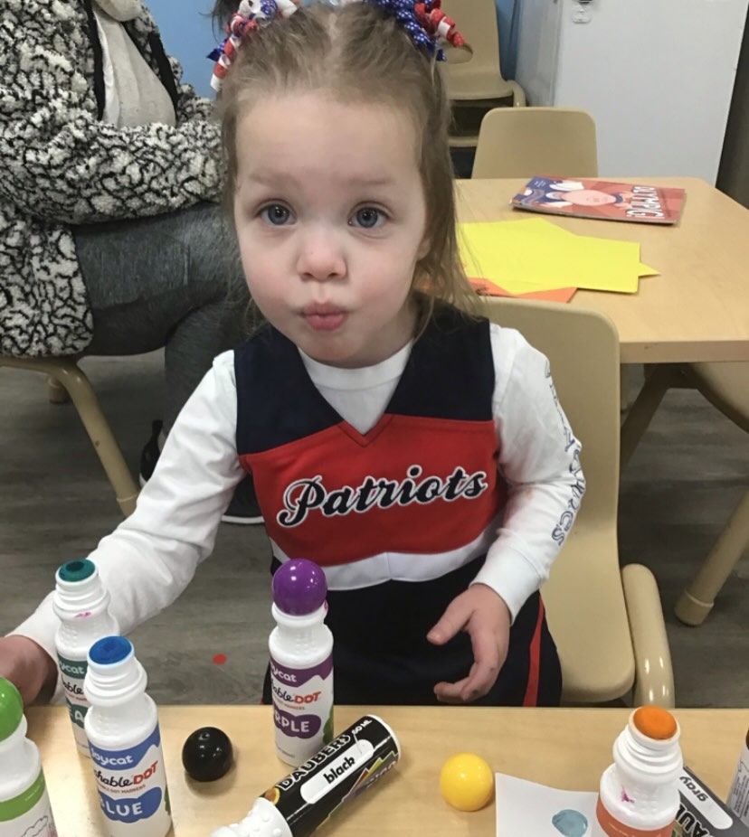 Child with Patriots shirt, making art. Pouting, surrounded by paint bottles, sitting at a small table.