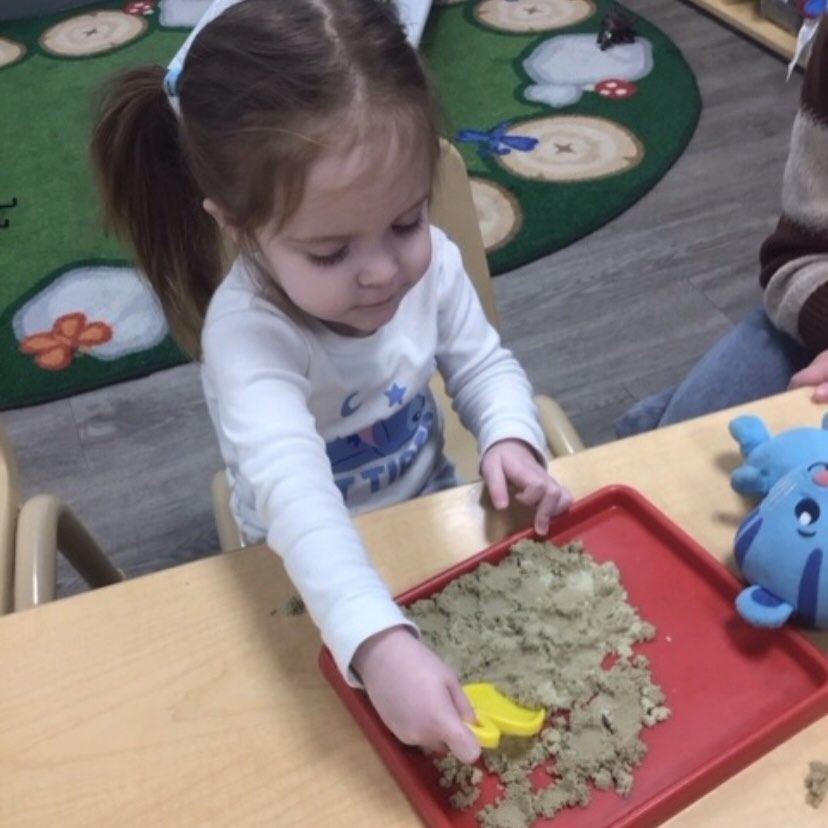 Young child plays with sand in a red tray at a table, using a yellow scoop.