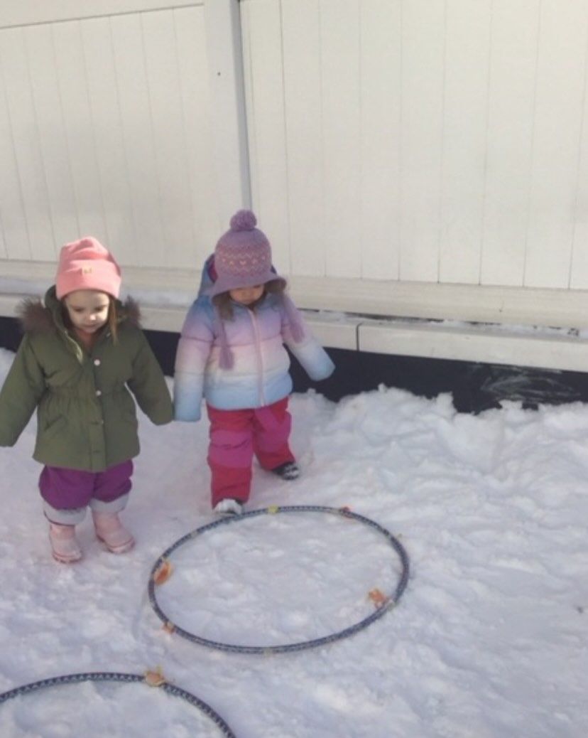 Two children in winter coats stand near hula hoops in the snow.