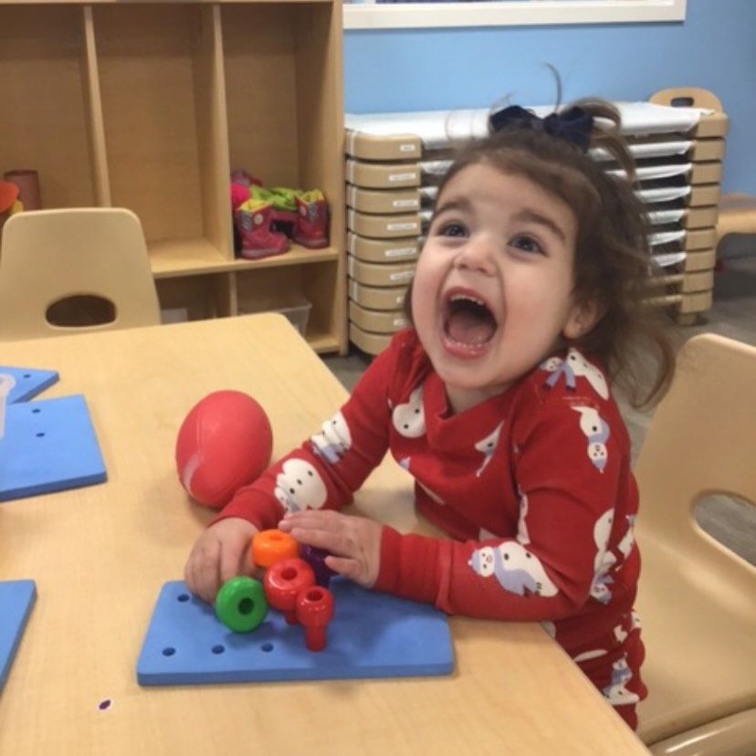 Child in red pajamas with bow, laughing at table with toys.