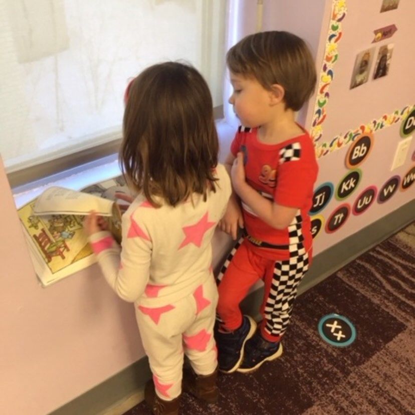 Two children in pajamas look at a book, by a window. One child wears pink stars, the other red racing attire.