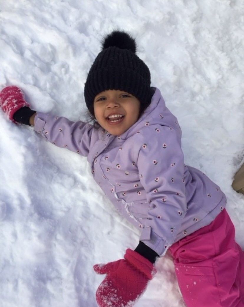 Child in snow, wearing black hat, pink coat and pants, smiling and lying in snow.