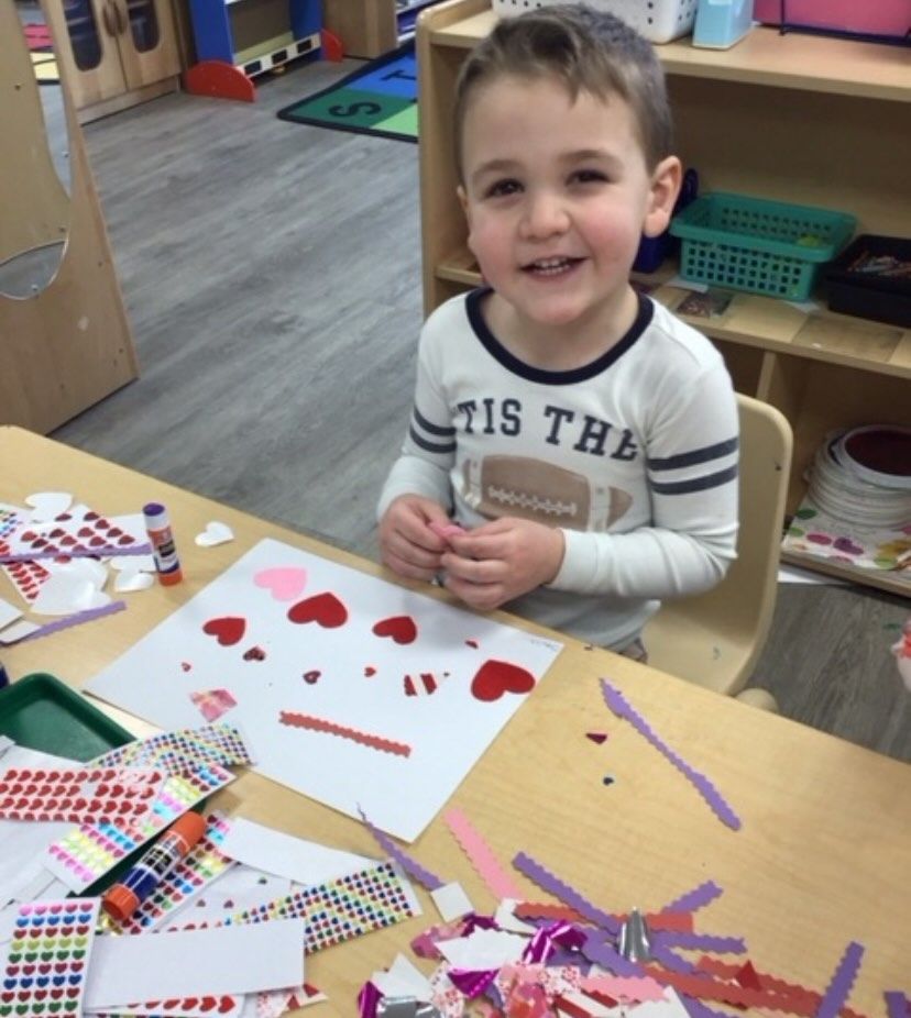 Boy smiles while crafting with heart-shaped stickers and paper at a desk.