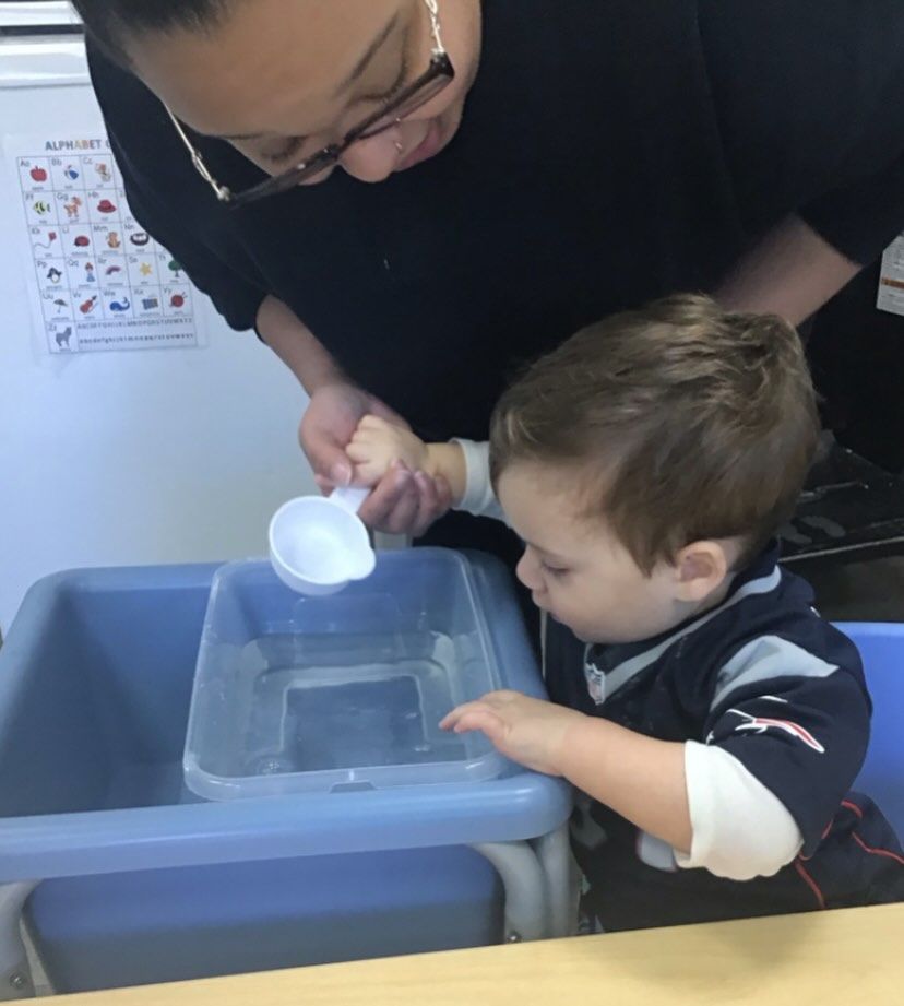 Adult helping a child pour water from a scoop into a basin. They are indoors.