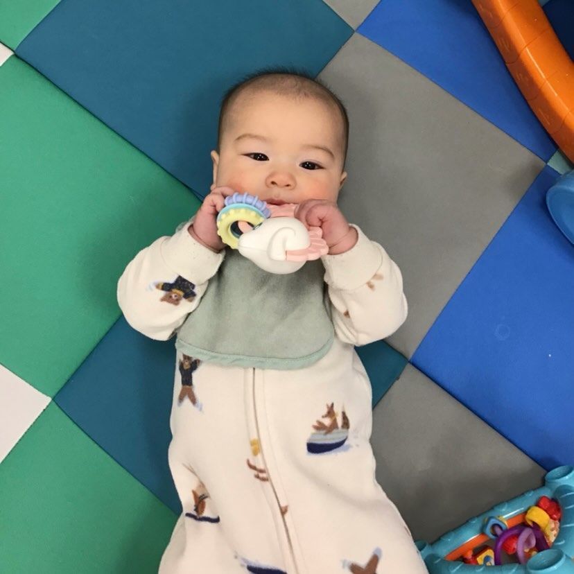 Baby in a onesie lying on a mat, chewing on a toy.