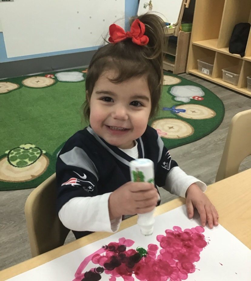 A child in a Patriots jersey smiles while using a paint marker at a table in a classroom setting.