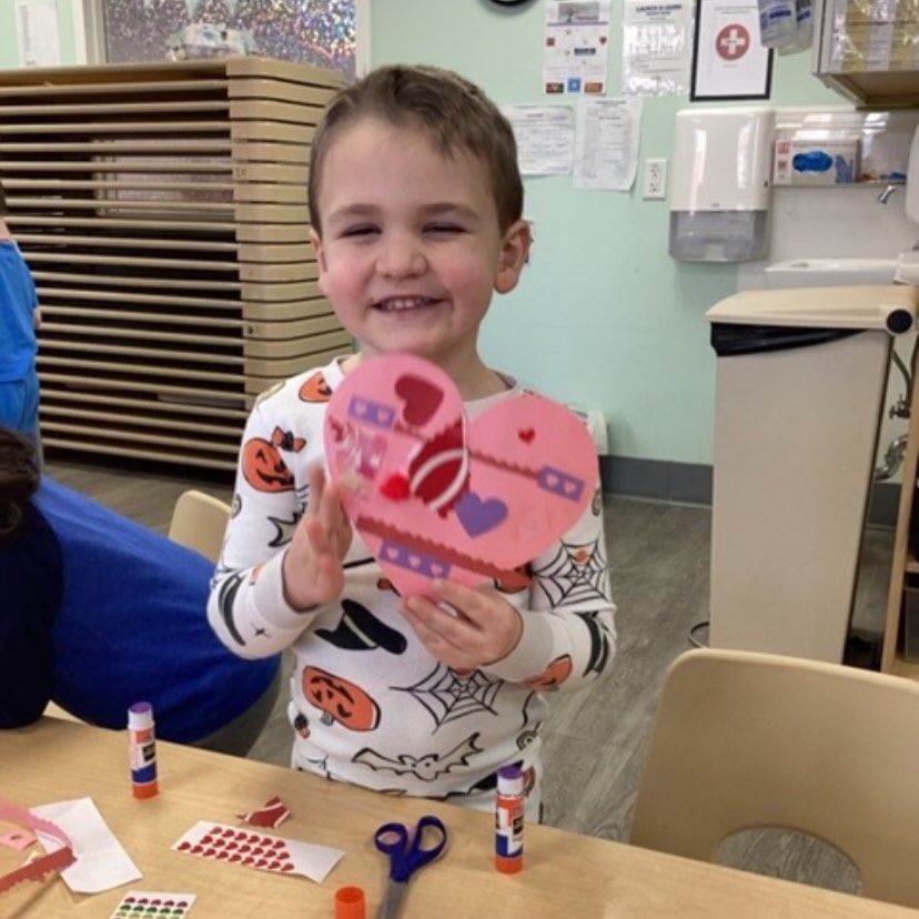 Boy holding a heart-shaped Valentine's Day craft, smiling at the camera. He wears pajamas and sits at a table.