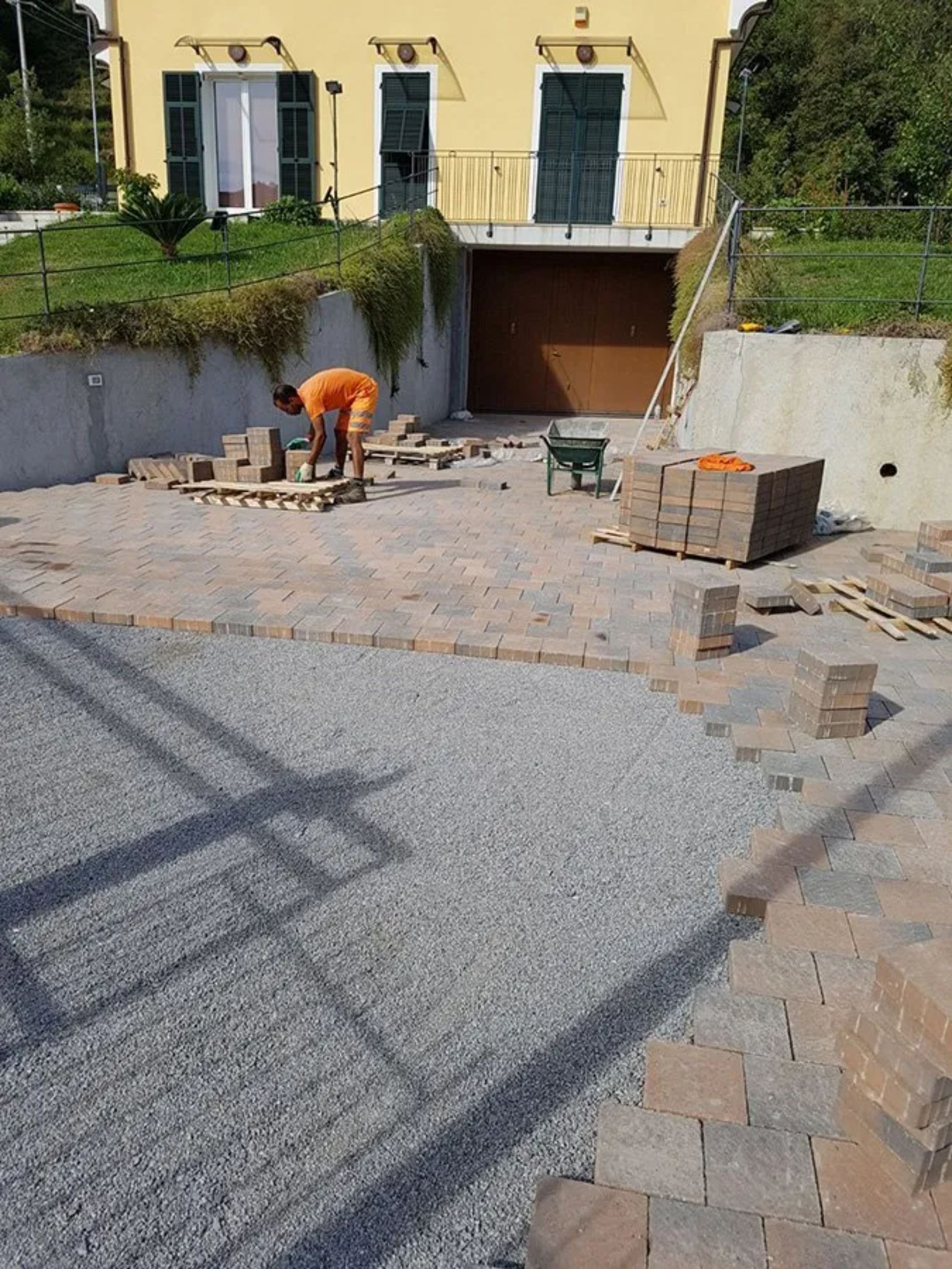 Un operaio edile posa la pavimentazione davanti a una casa gialla con garage. Sono visibili un sentiero di ghiaia, mattoni e una carriola.