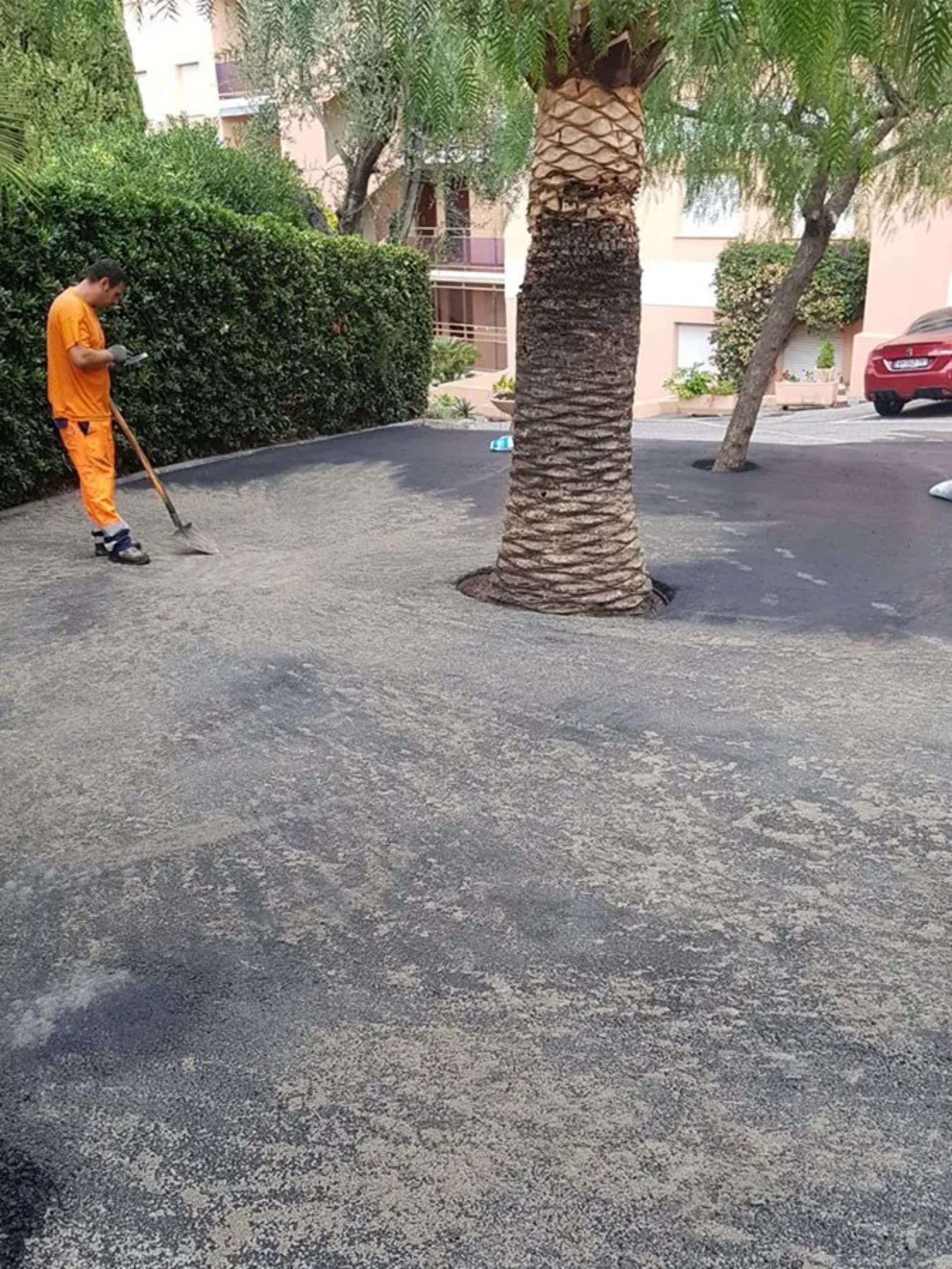 Un uomo in uniforme arancione rastrella la ghiaia su una superficie nera vicino a una palma e a dei cespugli verdi.