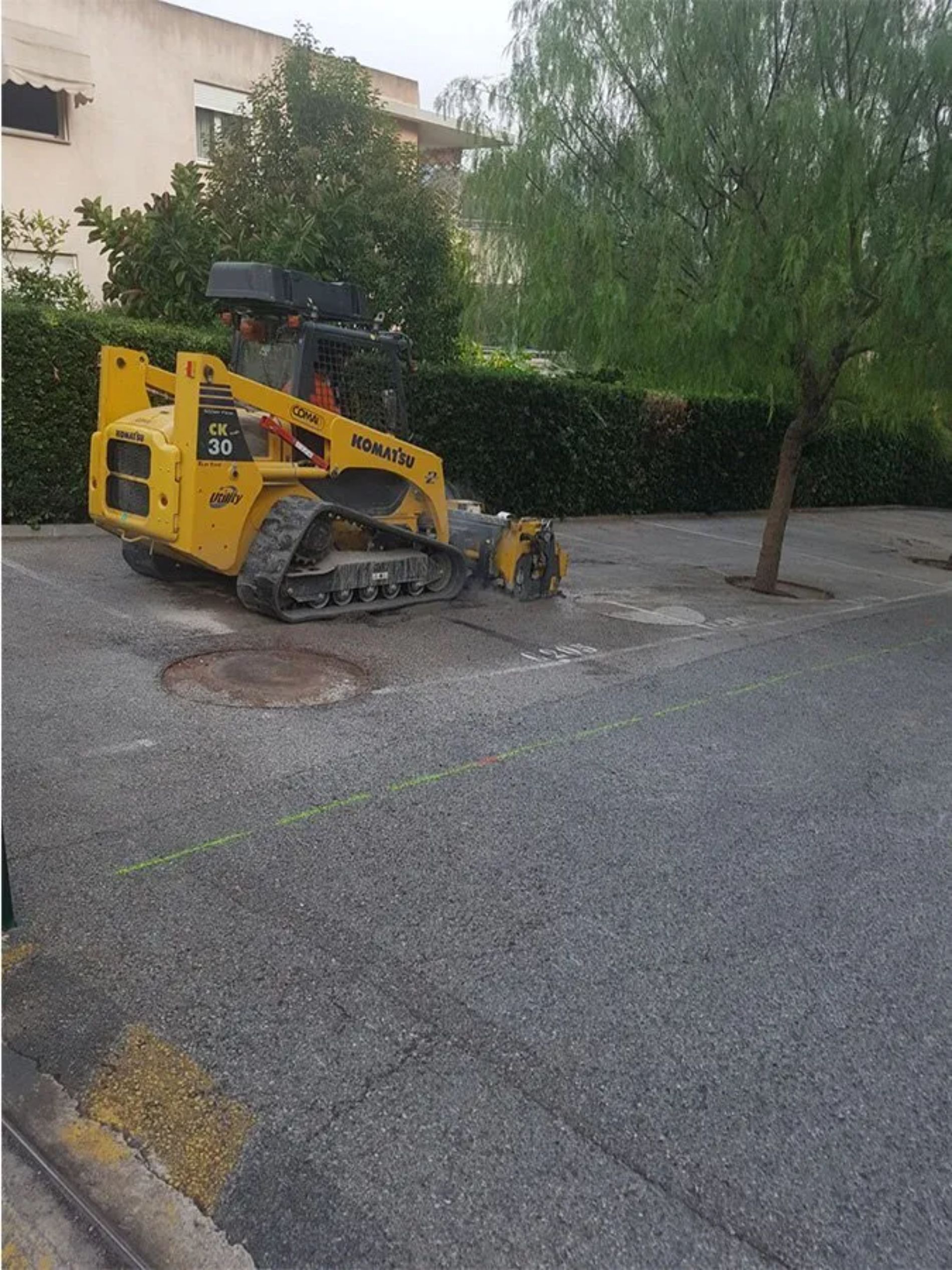 Minipala Komatsu gialla su asfalto, vicino a un albero e a un edificio, in fase di pavimentazione o riparazione.