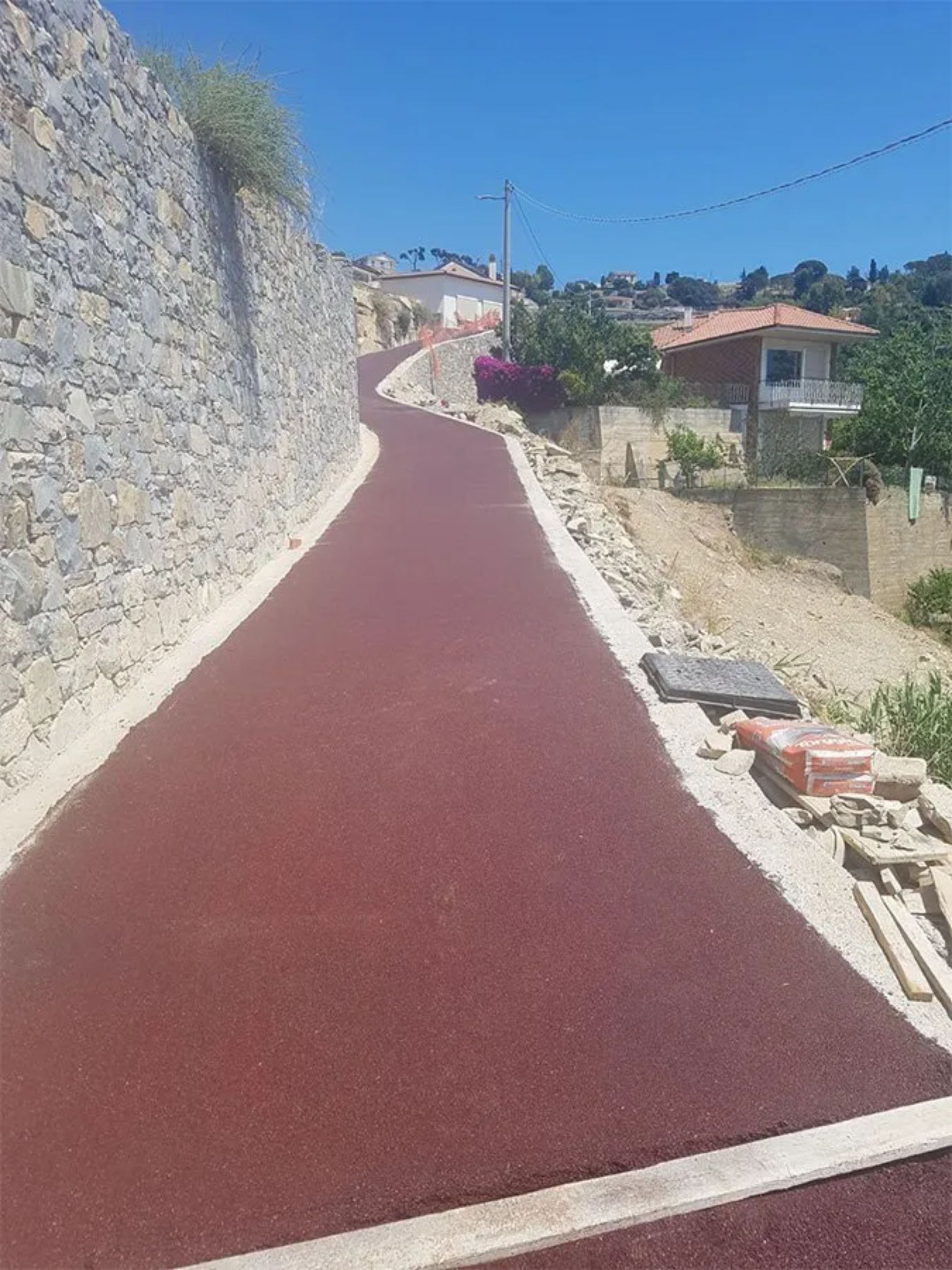 Una strada rossa, asfaltata e tortuosa in salita, con un muro di pietra da un lato e case/vegetazione dall'altro, sotto un cielo azzurro.