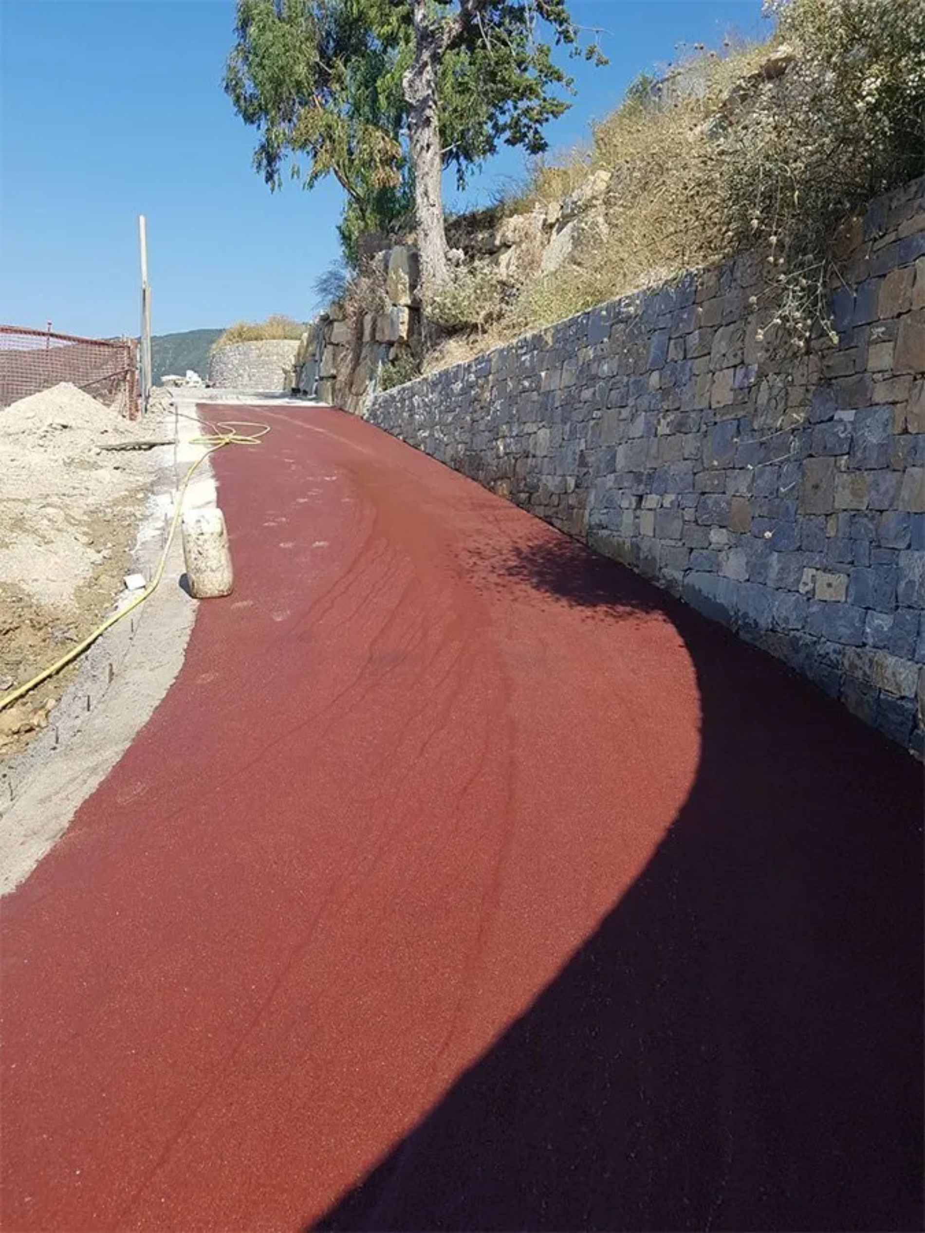 La strada asfaltata rossa sale, costeggiando un muro di pietra sul pendio di una collina. Giornata di sole.