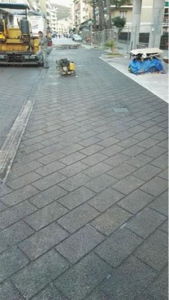 Operai che asfaltano una strada con lastre grigie simili a mattoni. Sullo sfondo si vedono una macchina e una persona.