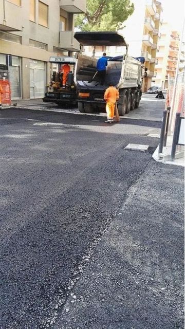 Pavimentazione stradale in corso. Operai in arancione e blu, camion dell'asfalto, edifici sullo sfondo.