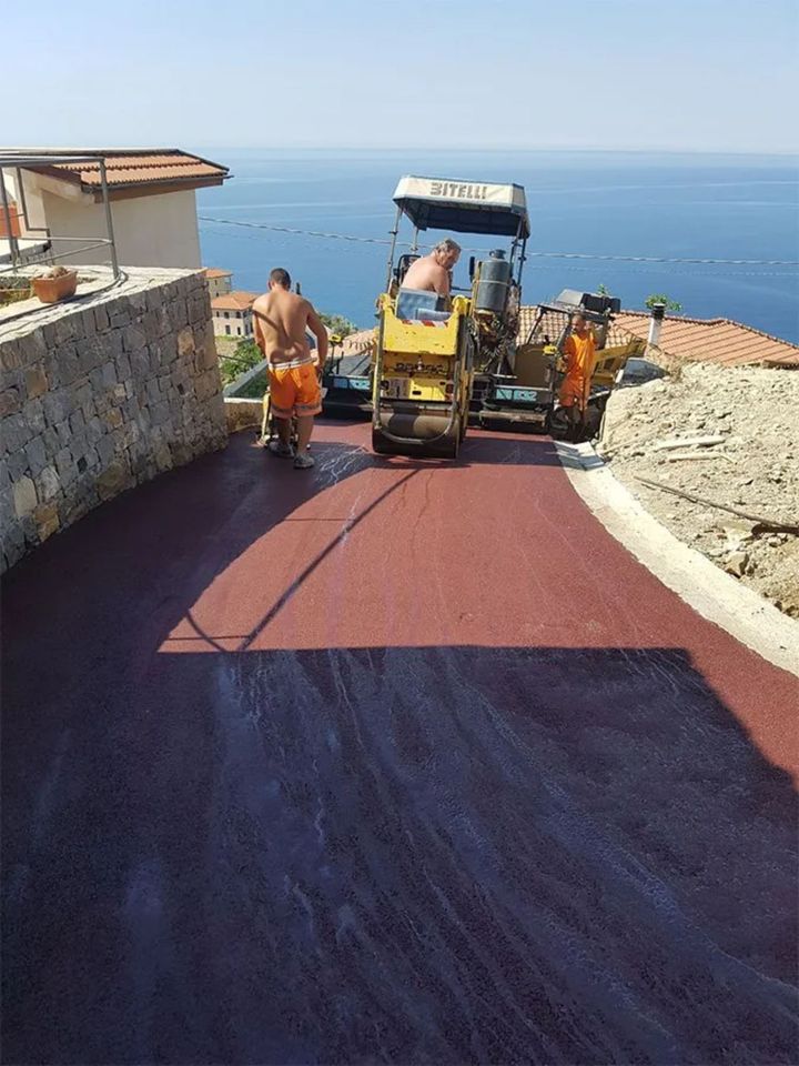Operai stradali che asfaltano una strada asfaltata rossa vicino a un panorama costiero in una giornata di sole.