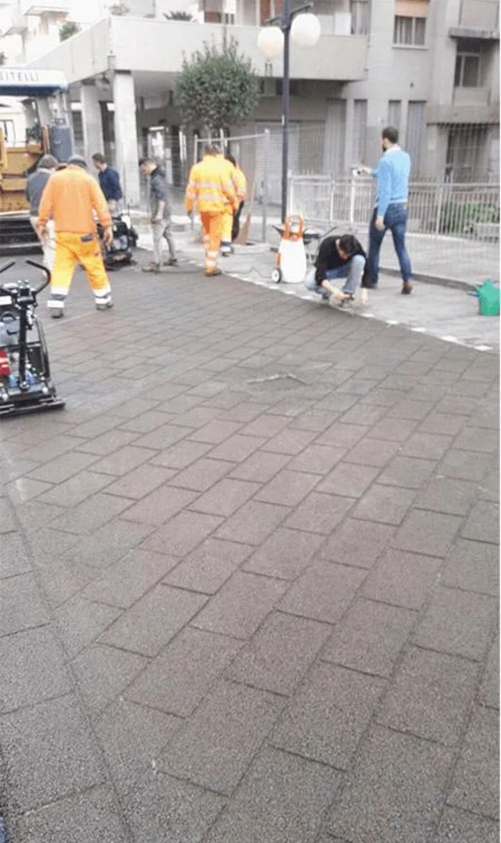 Lavoratori con giubbotti di sicurezza pavimentano uno spazio pubblico con piastrelle di gomma.