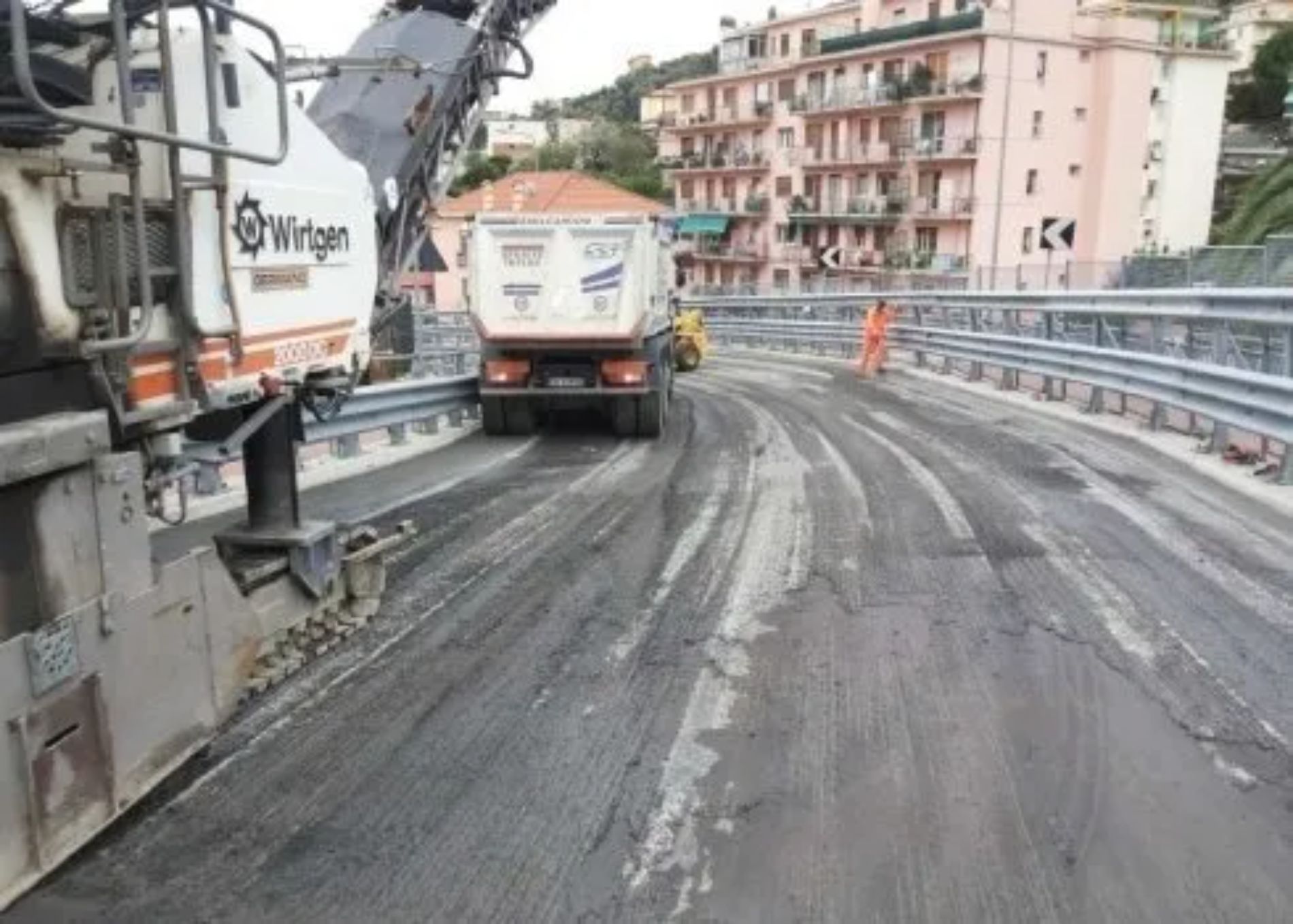 Una fresatrice stradale rimuove l'asfalto da un ponte, seguita da un autocarro con cassone ribaltabile.