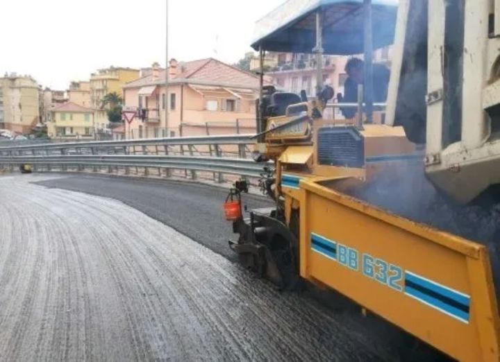 Una finitrice gialla stende l'asfalto su una strada curva. Gli edifici costeggiano la strada.