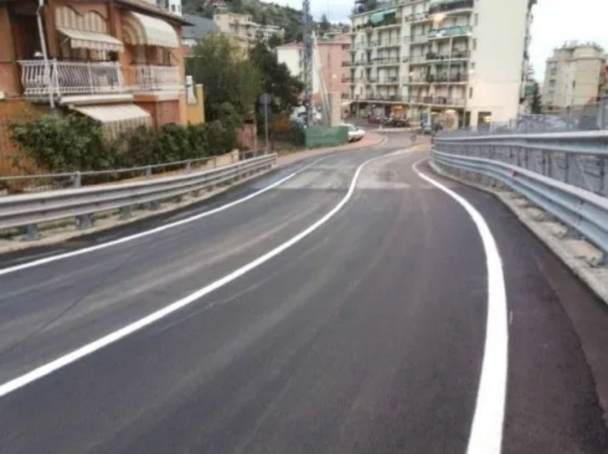 La strada curva verso l'alto, con segnaletica orizzontale bianca, guardrail metallici e edifici sullo sfondo.