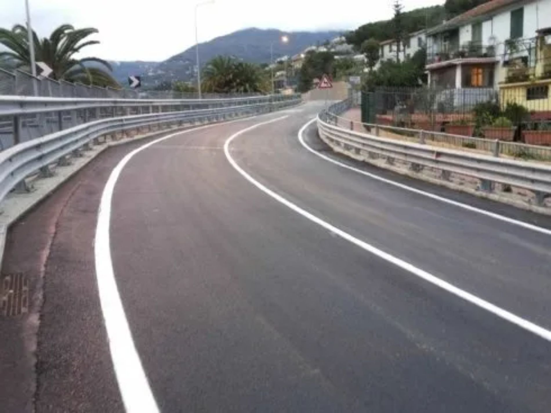 Strada asfaltata curva con segnaletica orizzontale bianca, fiancheggiata da guardrail, che sale verso una collina e alcuni edifici.