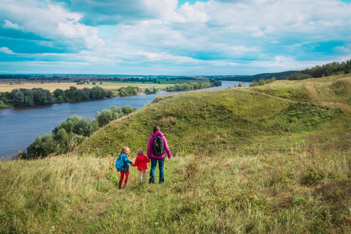 Person holding hands with two children, walking down a grassy hill overlooking a river.