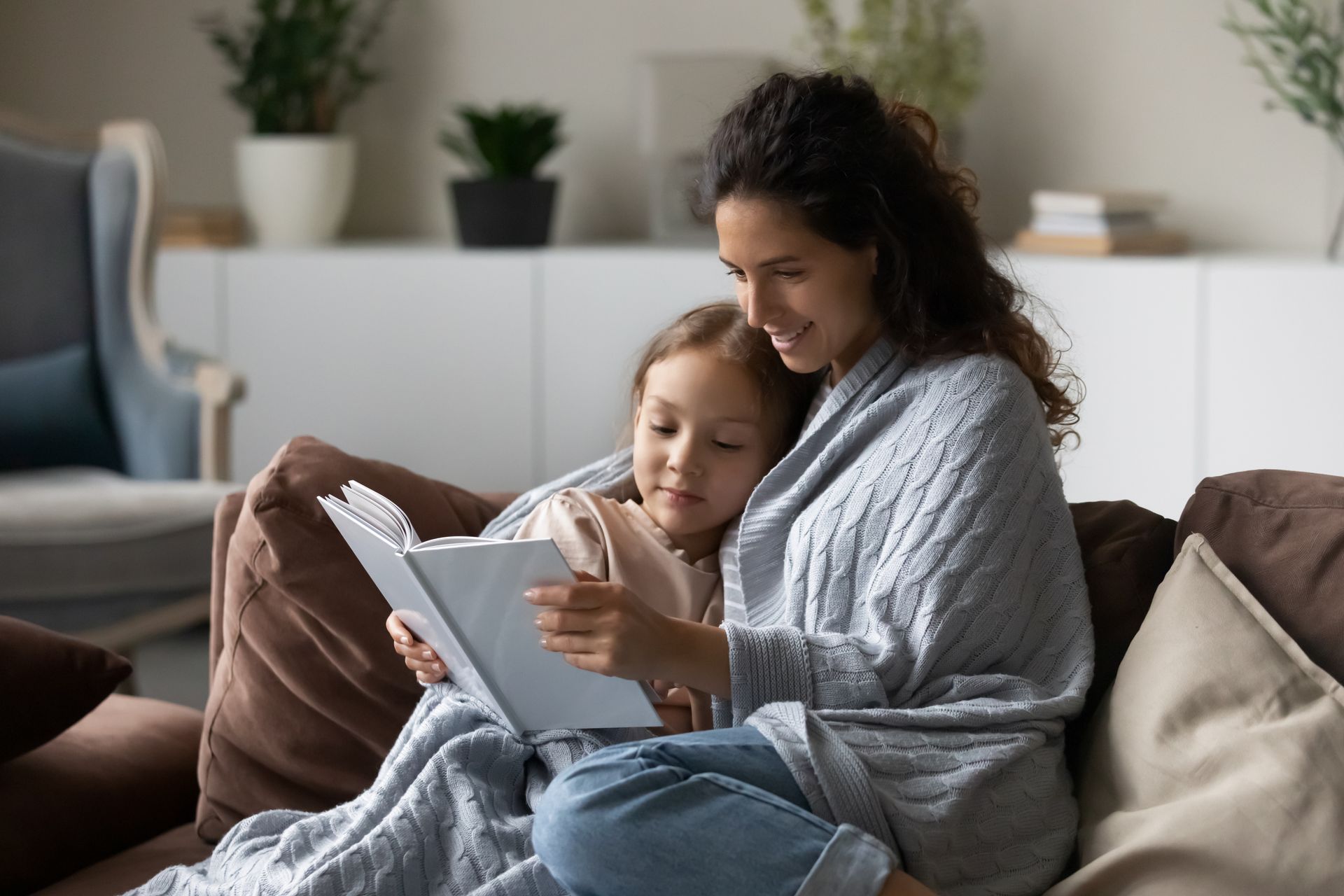 Woman and child reading a book together on a couch, wrapped in a blanket.