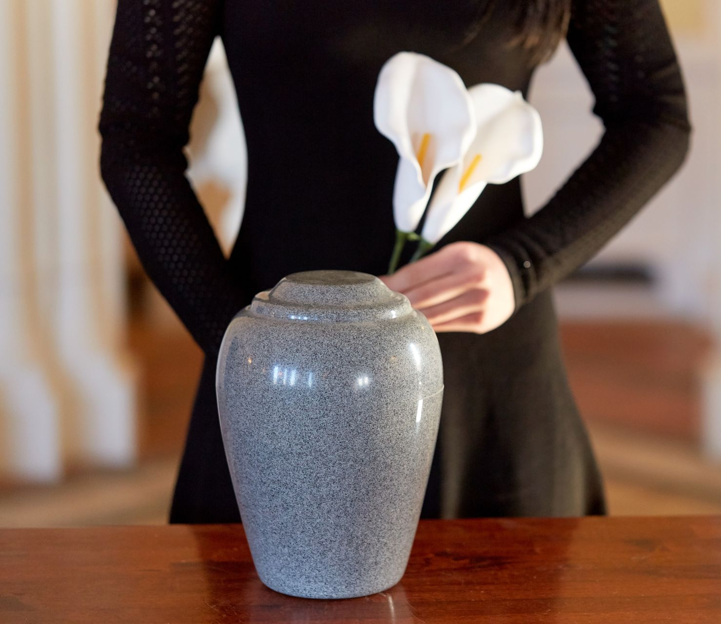 Person in black dress holding white calla lilies, stands near a granite-colored urn on a wooden table.