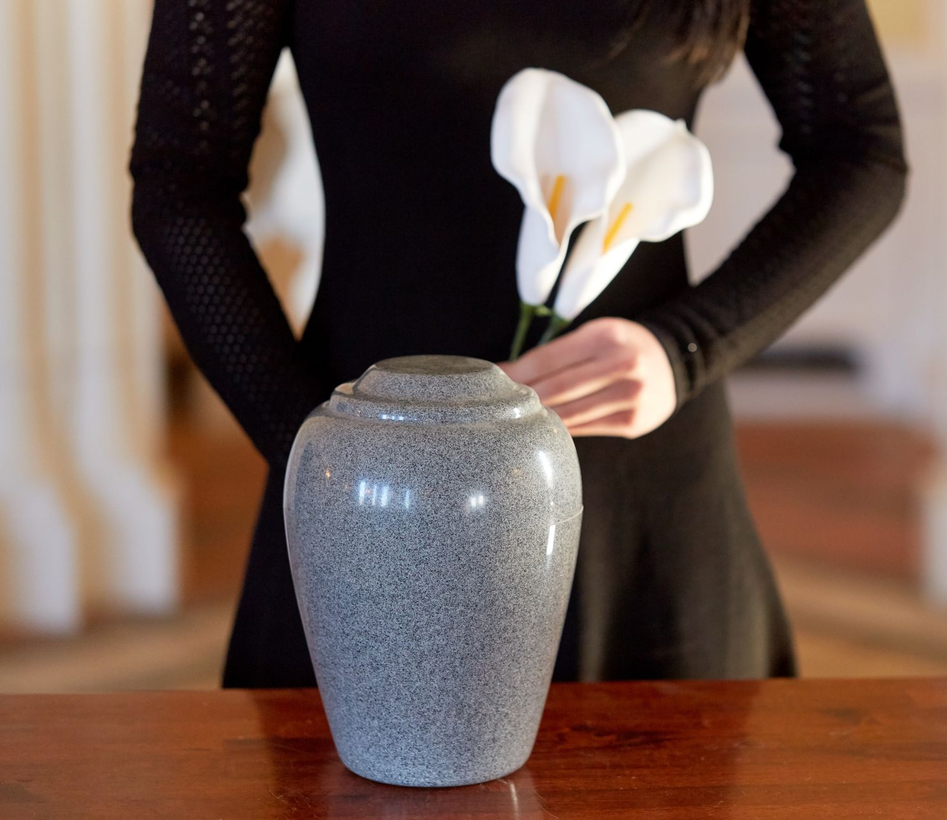 Person in black dress holding white calla lilies, stands near a granite-colored urn on a wooden table.