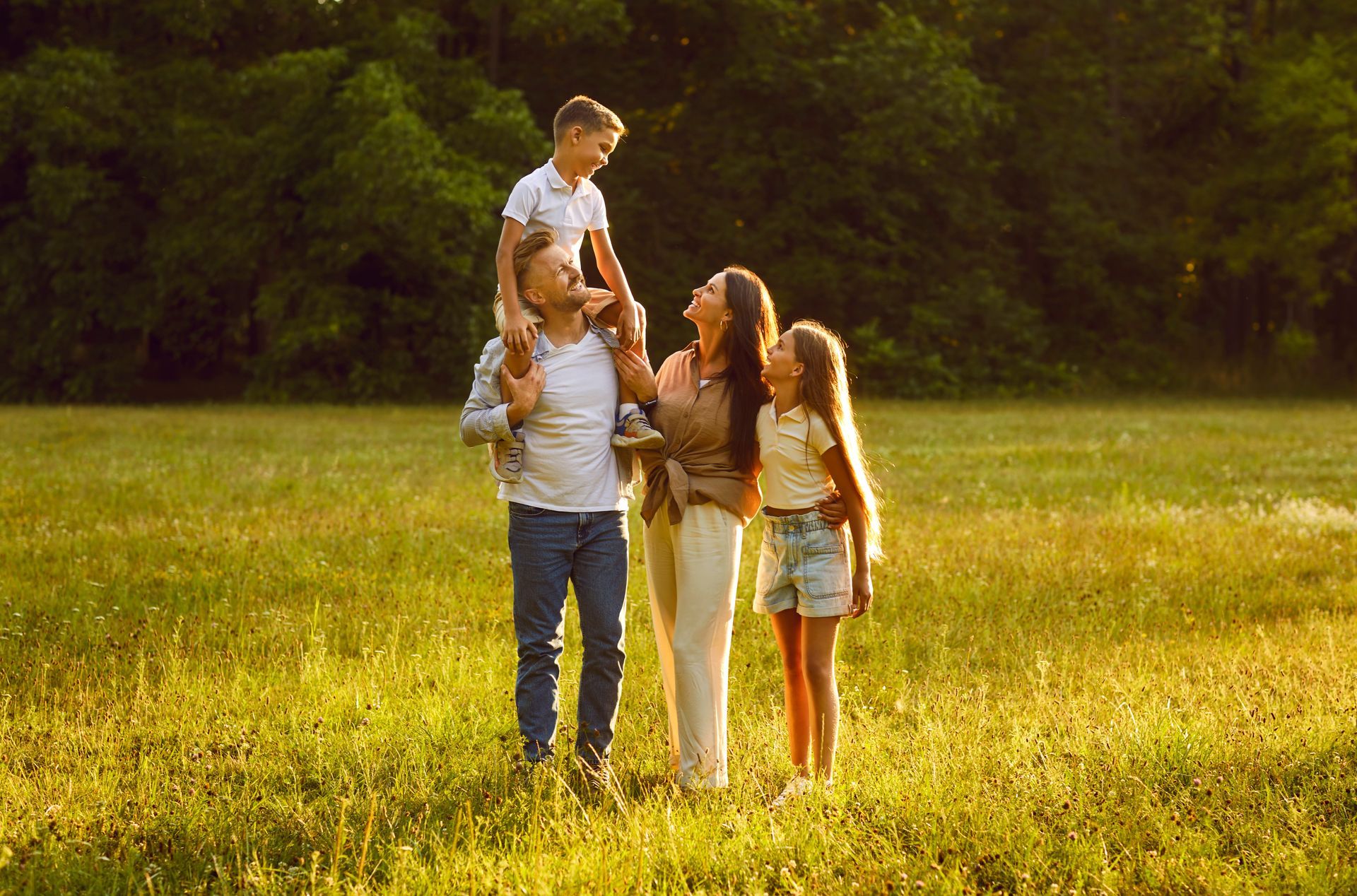 Family of four smiling in a grassy field; boy on shoulders, sunny lighting.