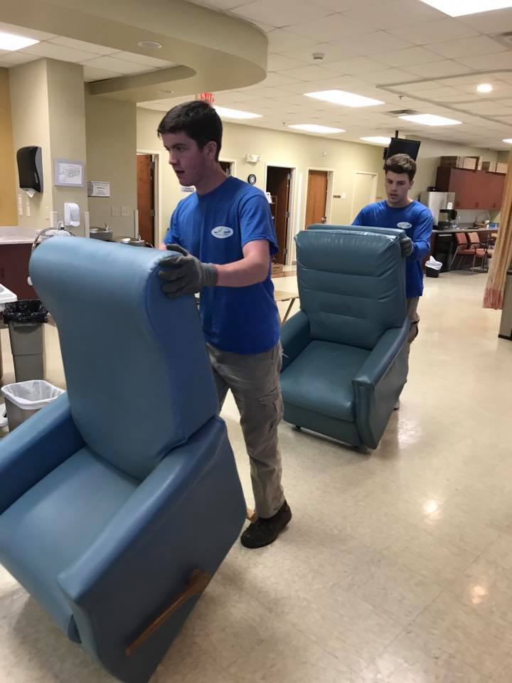 Two men are carrying recliner chairs in a hallway.