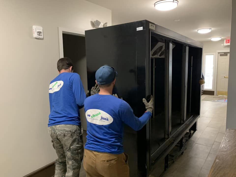 Two men are carrying a large black refrigerator in a hallway.