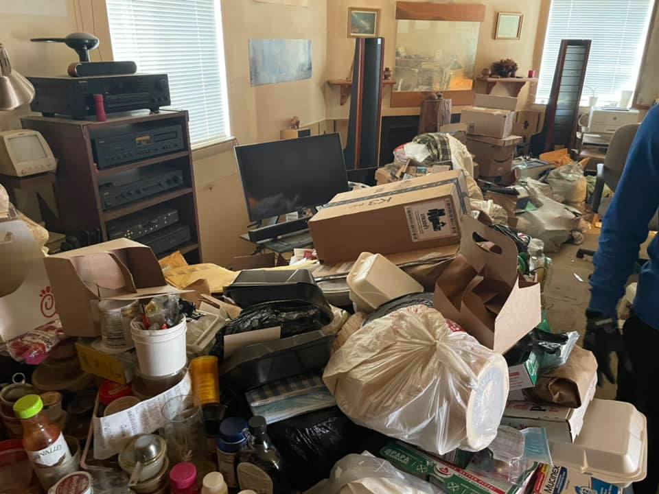 A person is standing in a living room filled with lots of trash.