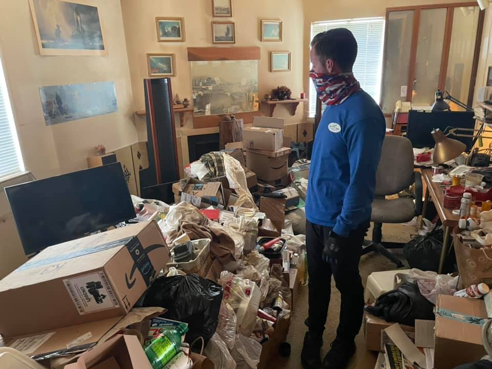 A man wearing a bandana is standing in a messy living room.