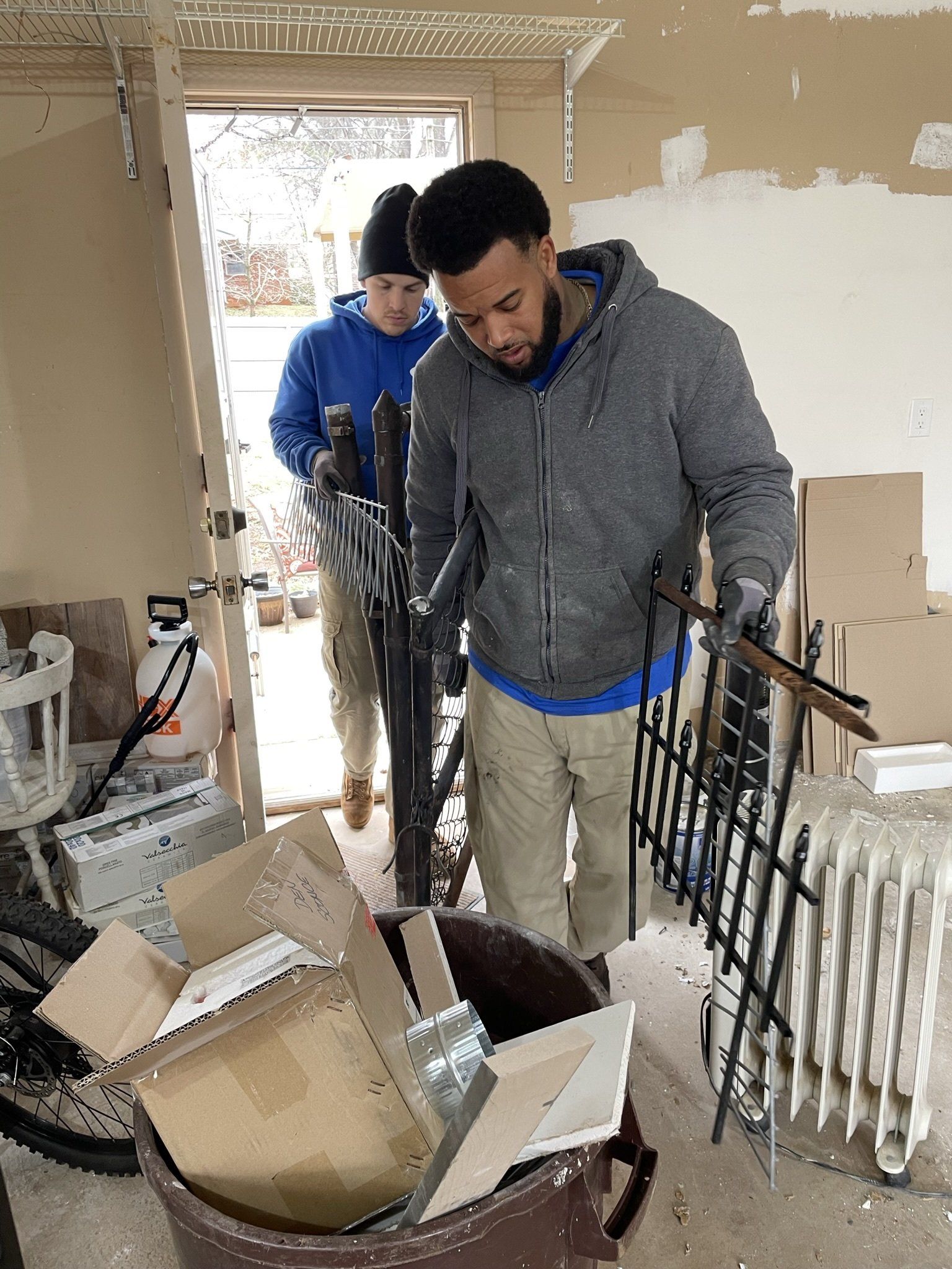 Two men are standing next to a bucket of junk in a room.