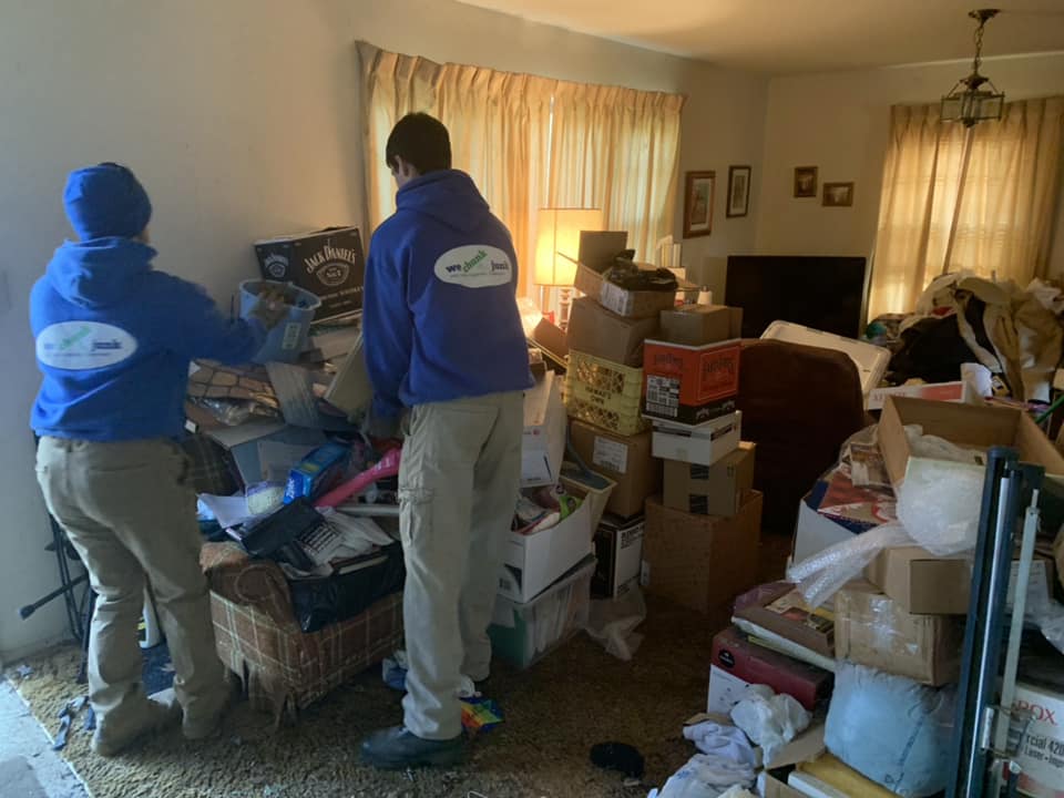 Two men are standing in a living room filled with boxes.