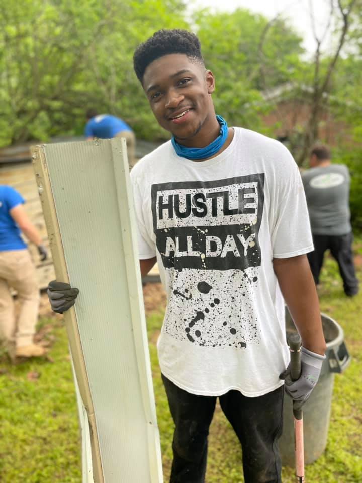 A young man wearing a white t-shirt that says `` hustle all day '' is holding a piece of wood.