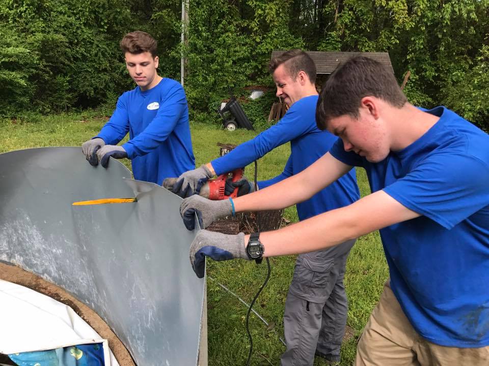 Three young men in blue shirts are working on a piece of metal