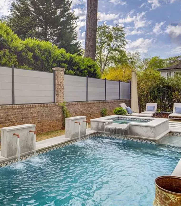 Swimming pool with water features, surrounded by a fence and lush greenery, under a cloudy sky.