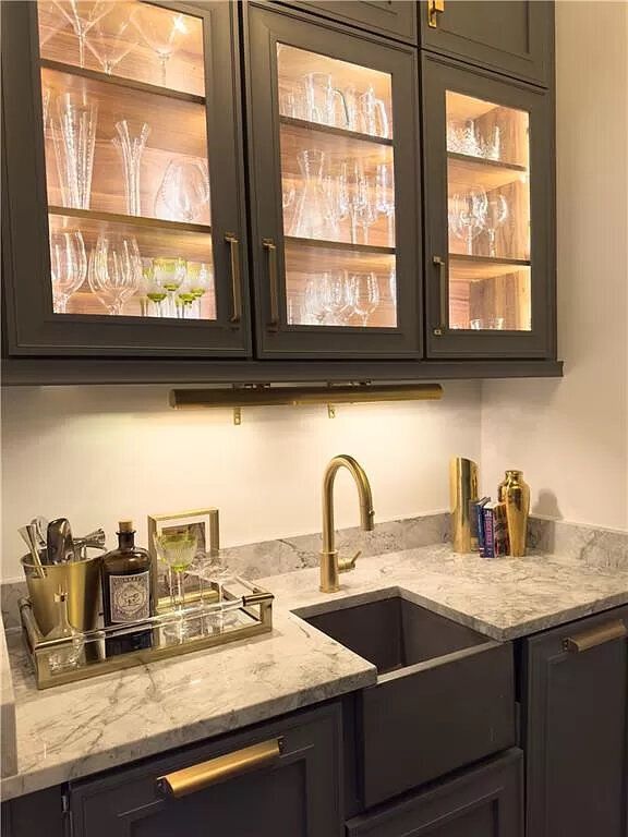 Dark cabinets with illuminated glass shelves display glassware above a sink with a gold faucet.